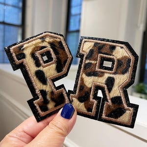 May include: Two embroidered letter patches, the letter "R", in a leopard print design. The patches have a black border with a brown trim. The background is a neutral color.