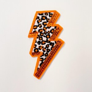 May include: Orange and black leopard print lightning bolt iron-on patch. The patch is made of sequins and is perfect for adding a touch of personality to clothing, bags, or other accessories.