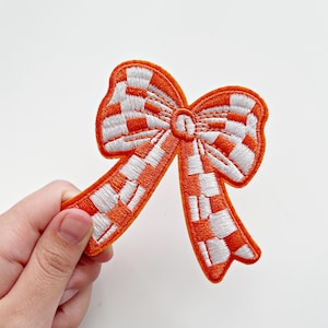 May include: Embroidered orange and white checkered bow patch. The bow has a detailed design with a bright orange outline and a checkered pattern in white and orange. This patch is perfect for adding a decorative touch to clothing or accessories.
