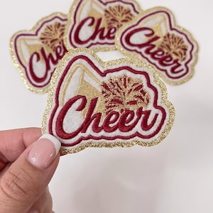 May include: A set of embroidered patches featuring the word Cheer in dark red script, accompanied by a white megaphone and a dark red pom-pom graphic. Each patch has a white background and a shimmering gold glitter border, creating a distinct outline.