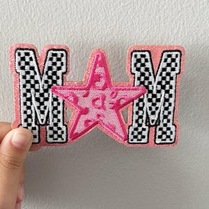 May include: Pink embroidered patch with the letters "M A M" and a star in the center. The letters have a black and white checkered pattern, and the star has a pink floral design.