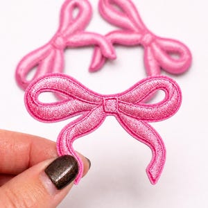 May include: Three bright pink embroidered bow patches with a satin-like sheen and stitched edges. Each patch features a central knot with two loops and two downward-curving tails, showcasing a detailed thread texture throughout the design.