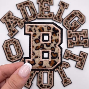 May include: A collection of varsity-style letter patches featuring a brown and black leopard print pattern. Each patch has a thick black border and a light pink stitched outline. The letter B is held in the foreground, with other letters visible.