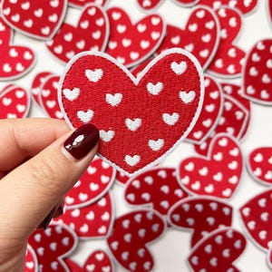 May include: Red heart-shaped patches with white heart patterns. The patches are embroidered with a white border. One patch is held up, with many more scattered in the background. The patches are suitable for crafts and DIY projects.