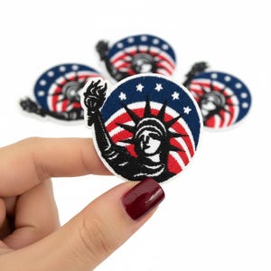 May include: A circular embroidered patch featuring the Statue of Liberty in black thread against a background of red and white stripes and a blue border with white stars. The patch is held up, with three additional identical patches visible behind it.