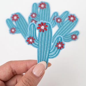 May include: A light blue embroidered cactus patch featuring vertical ribbed detailing and several small red flowers with white centers. The cactus has a central stem with two curved side arms, each adorned with red and white floral accents.