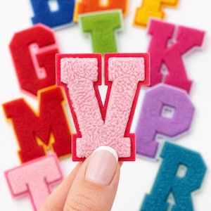 May include: A collection of colorful, fuzzy, varsity-style letter patches in various shades including blue, orange, yellow, red, green, pink, and purple. A light pink letter V patch with a textured surface is held in the foreground.