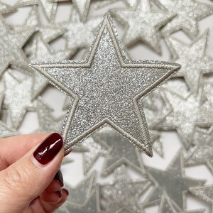 May include: A silver glitter star with a white outline. The star is about 3 inches wide and is surrounded by other silver glitter stars.