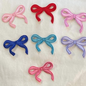 May include: Assortment of eight embroidered iron-on patches in various colors, featuring a bow design. The bows are in pink, red, blue, light blue, purple, and lavender.