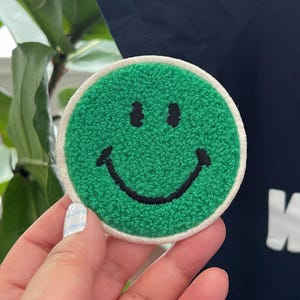 May include: Green fuzzy smiley face patch with a white border. The patch is held in a hand.