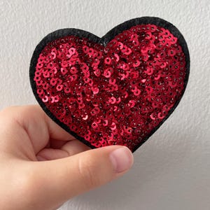 May include: A red sequined heart-shaped patch with a black felt border. The heart is filled with shiny red sequins. The patch is held up by a hand against a white background.