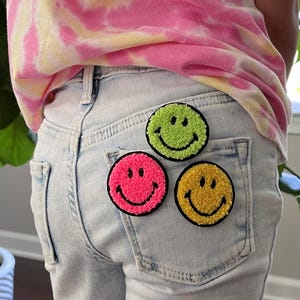 May include: Three embroidered smiley face patches in different colors: green, pink, and yellow. The patches are sewn onto the back pocket of a pair of light blue denim shorts.