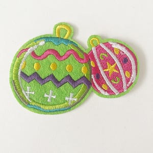 May include: Two embroidered Christmas ornament patches. One is green with a zig-zag pattern, yellow dots, and white crosses. The other is pink with white stripes and a yellow star. Both have yellow loops.