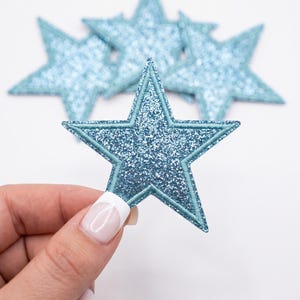 May include: A set of light blue star-shaped patches featuring a glittery texture and a stitched border. The patches are five-pointed stars with a shimmering finish, designed for application to fabric surfaces with a clear, defined edge.