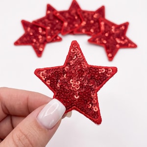 May include: A collection of five star-shaped patches, each covered in vibrant red sequins. The patches feature a stitched red border around the edges. One individual star patch is held in the foreground, while four additional identical patches are arranged behind it.