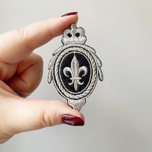 May include: A silver embroidered patch with a black background featuring a fleur-de-lis design and a crown at the top.