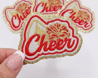 Könnte beinhalten: Ein Set aus gestickten Aufnähern mit dem Wort Cheer in roter Schreibschrift, begleitet von einer Grafik eines goldenen Megafons und Pompons. Jeder Aufnäher hat einen weißen Hintergrund, einen roten Rand und eine goldene Glitzerkante.