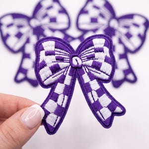 May include: An embroidered patch in the shape of a bow featuring a purple and white checkered pattern. The patch has a thick purple border and a central knot detail. Two additional identical bow patches are visible in the blurred background.