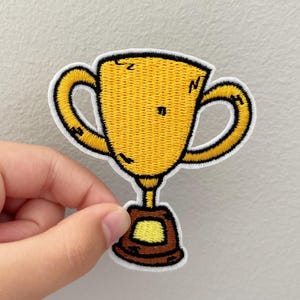 May include: Embroidered patch featuring a yellow trophy with black outlines and a white border. The trophy has two curved handles and a brown base with a yellow square. The patch is held against a light gray background.