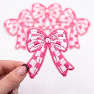 May include: A set of embroidered iron-on patches shaped like bows, featuring a pink and white checkered pattern. Each bow has a distinct pink border and a central knot detail. The patches are arranged with one in the foreground and three behind.