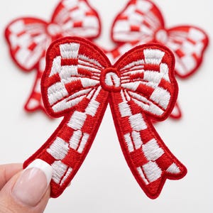 May include: Three embroidered bow patches featuring a red and white checkered pattern. Each bow has a distinct knot in the center and two flowing tails. The embroidery is dense, creating a textured surface with a clear contrast between the colors.
