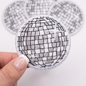May include: A circular embroidered patch featuring a disco ball design with alternating silver glitter and grey square segments. The patch has a white stitched border and includes two white starburst accents on the surface. Three additional identical patches are visible in the background.