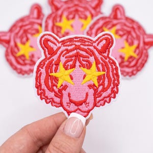 May include: An embroidered patch featuring a pink tiger head with red stripes and bright yellow stars covering the eyes. The patch has a white border and is shown with several identical patches arranged in the background.