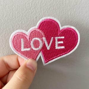 May include: Embroidered patch featuring two overlapping hearts in shades of pink. The word "LOVE" is stitched in white across the larger heart. The patch has a white border and is likely for clothing or accessories.