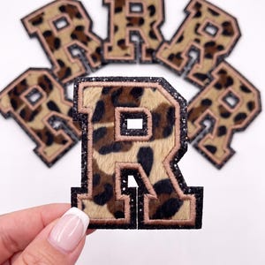 May include: A collection of letter R patches featuring a brown and black leopard print pattern. Each patch has a glittery black border and a light pink stitched outline. The letter R is designed in a block varsity font style with a fuzzy texture.