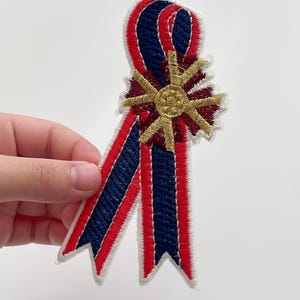 May include: A red, white, and blue embroidered ribbon with a gold star in the center. The ribbon is folded into a bow shape.