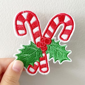 May include: Embroidered Christmas patch featuring two red and white striped candy canes, green holly leaves, and red berries. The patch has a white border and is designed for sewing or ironing onto fabric. A festive holiday decoration.