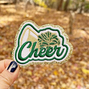 May include: Green and gold glitter iron-on patch with the word "Cheer" in a white outline. The patch features a green and gold cheerleader's pom-pom.