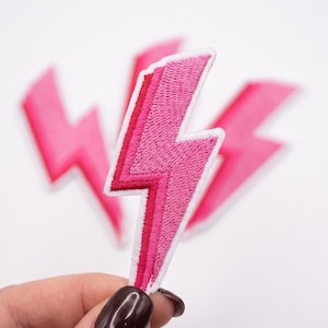 May include: A set of embroidered lightning bolt patches featuring a two-tone pink design with a darker pink center stripe and a lighter pink outer section, all outlined with a white border. The patches are shaped like jagged bolts of electricity.