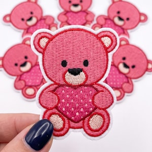 May include: A collection of embroidered iron-on patches featuring a pink teddy bear holding a pink heart with white polka dots. Each bear has a dark pink nose, black eyes, and light pink accents on the ears and feet, finished with a white border.