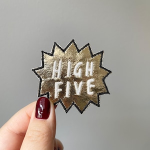 May include: Gold-colored fabric patch with a black outline in the shape of a starburst. The patch says "HIGH FIVE" in white lettering.