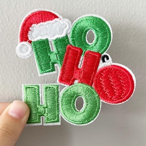 May include: Embroidered Christmas patches spelling "HO HO" in red and green. One patch features a Santa hat, and another includes a red ornament. The patches are held by a hand against a neutral background. Festive holiday decor.