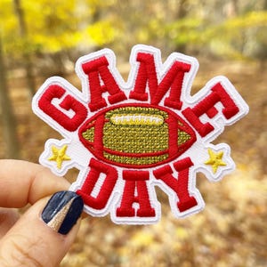 May include: Red and white embroidered patch with the words "GAME DAY" and a gold football in the center. The patch has two gold stars on either side of the football.