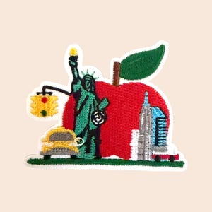 May include: An embroidered patch featuring a large red apple with a green leaf, the Statue of Liberty in teal, a yellow taxi cab, a traffic light, and a grey skyscraper. The design is set against a solid light background with a white border.