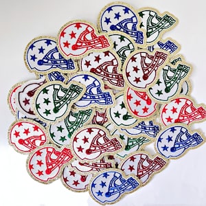 May include: A collection of iron-on patches featuring a football helmet with stars in various colors, including red, blue, green, and white. The patches have a gold glitter outline.