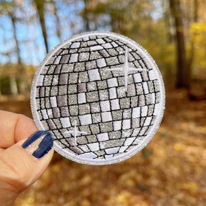 May include: A silver glitter disco ball patch with a white outline. The patch is round and has a textured surface.
