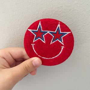 May include: A red, circular patch with a smiling face design. The face features two star-shaped eyes with blue and white accents and a white stitched smile. The patch is held by a hand.