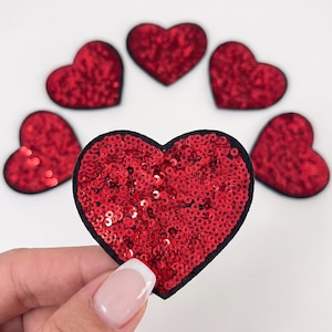 May include: A collection of six heart-shaped patches, each covered in small, reflective red sequins with a thin black border. One patch is held in the foreground, while five identical heart patches are arranged in a semi-circle behind it.