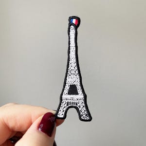 May include: A white embroidered iron-on patch of the Eiffel Tower with a small French flag on top. The patch is black outlined.