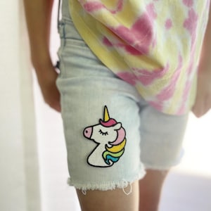 May include: Light blue denim shorts with a rainbow unicorn patch on the front. The shorts have a frayed hem.