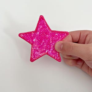 May include: A pink star-shaped patch covered in iridescent sequins. The patch is made of fabric and has a stitched edge.