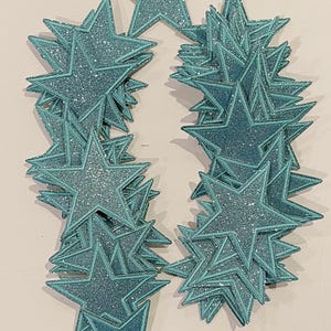 May include: A large stack of teal glitter star-shaped embellishments. The stars have a white outline and are perfect for crafting projects.