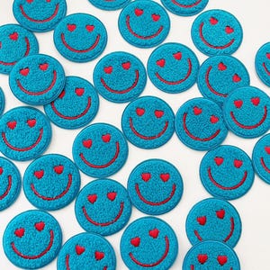 May include: A collection of teal-colored, round patches with a smiley face design. Each patch features a red embroidered smile and two red heart-shaped eyes. The patches are arranged on a white surface.