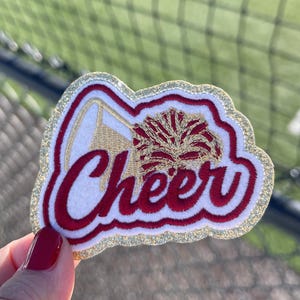 May include: A white felt patch with a gold glitter border. The patch features a red and gold embroidered cheerleader megaphone and pom pom with the word "Cheer" in red embroidery.
