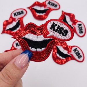 May include: A collection of red sequined lip-shaped patches featuring white teeth and a black mouth interior. Each patch includes a white speech bubble with the word KISS embroidered in black. The sequins provide a shiny texture to the lips.