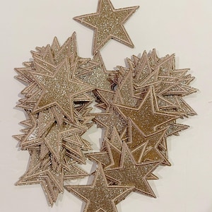 May include: A pile of gold glitter star-shaped iron-on patches. The patches are approximately 2 inches in diameter.